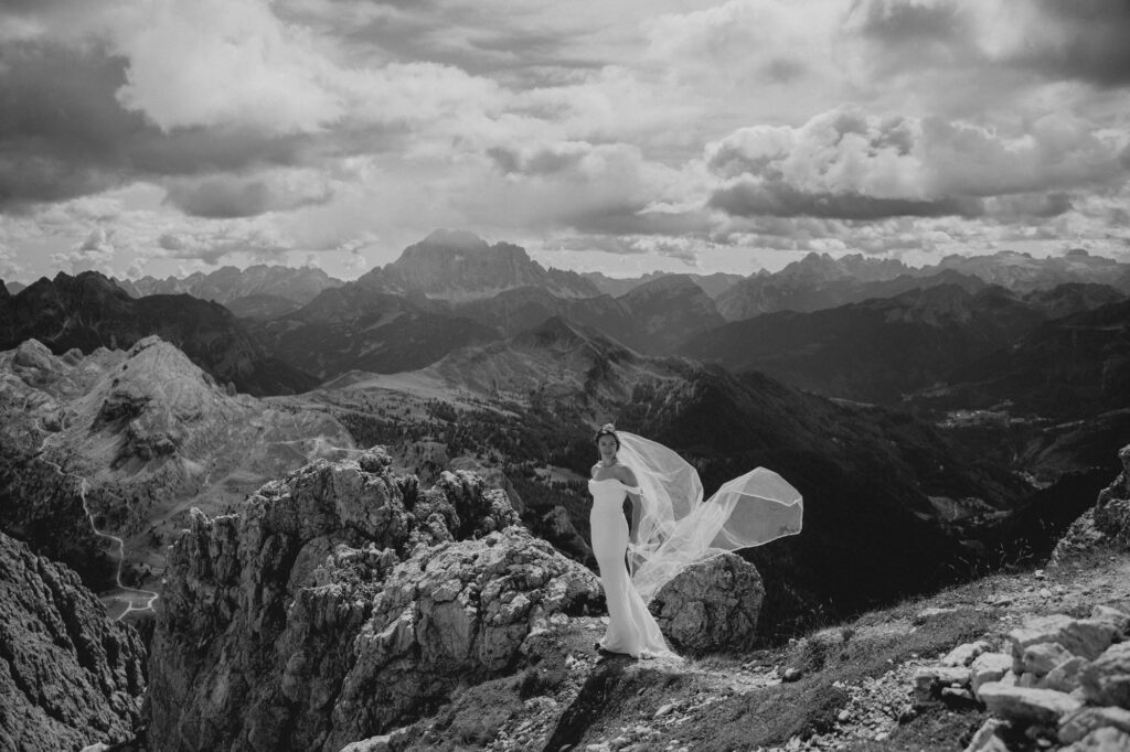 Braut im Hochzeitskleid steht bei einem Elopement in den Dolomiten allein auf einem Felsvorsprung, umgeben von dramatischer Berglandschaft, Schwarzweissaufnahme.