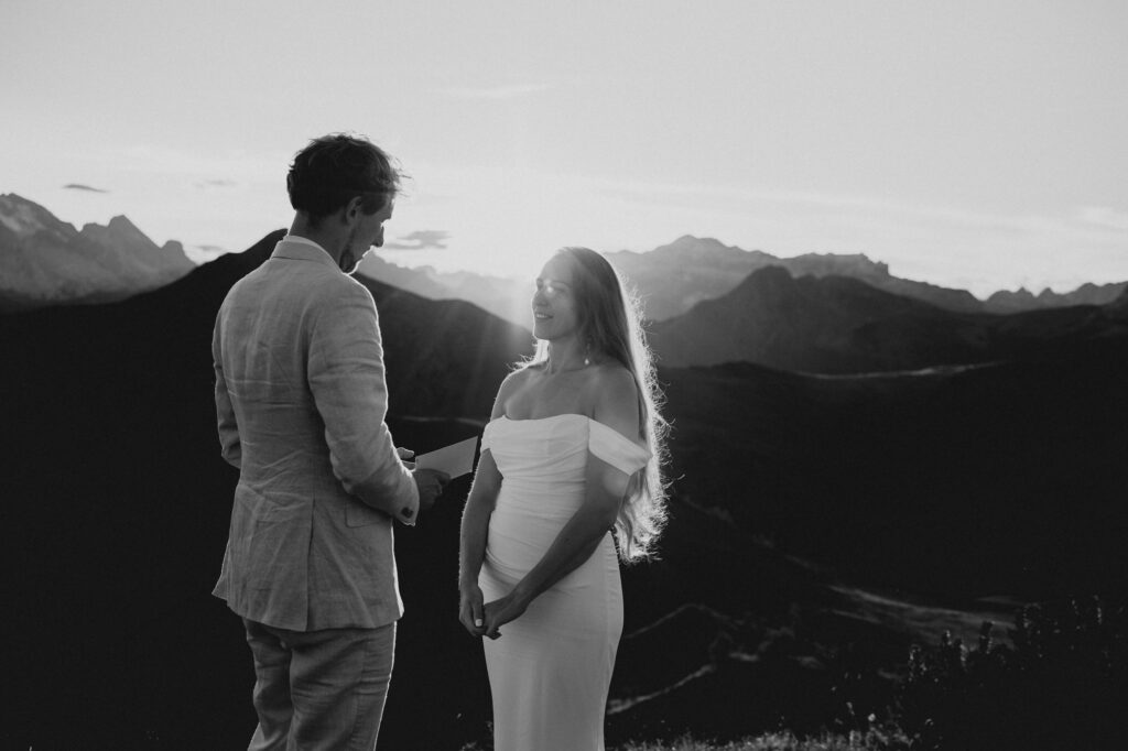 Braut und Bräutigam stehen bei einem Elopement in den Dolomiten Hand in Hand und blicken in die weite Berglandschaft bei Sonnenuntergang, Schwarzweissaufnahme.